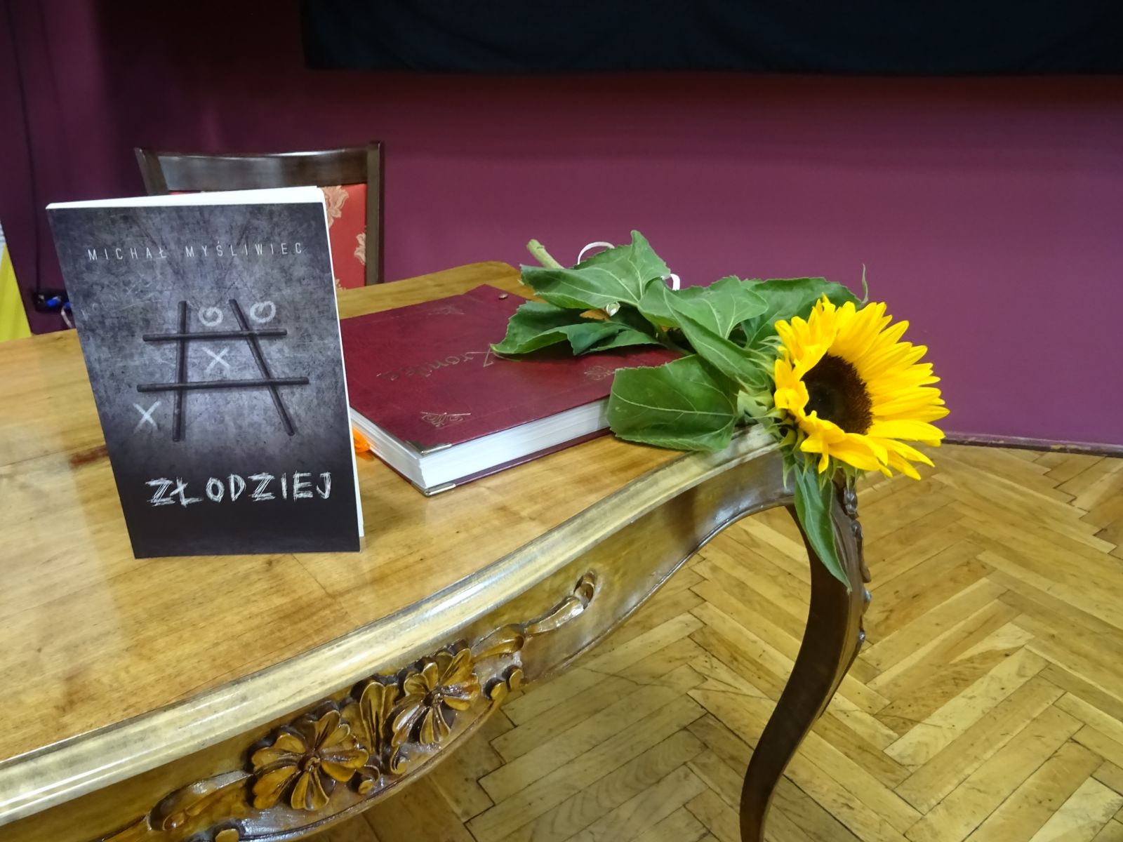 Fragment starego drewnianego stołu z rzeźbionym motywem kwiatowym z boku blatu. Na stole ustawiona promowana książka Złodziej, obok leży kronika biblioteki w czerwonej okładce i pojedynczy kwiat słonecznika.