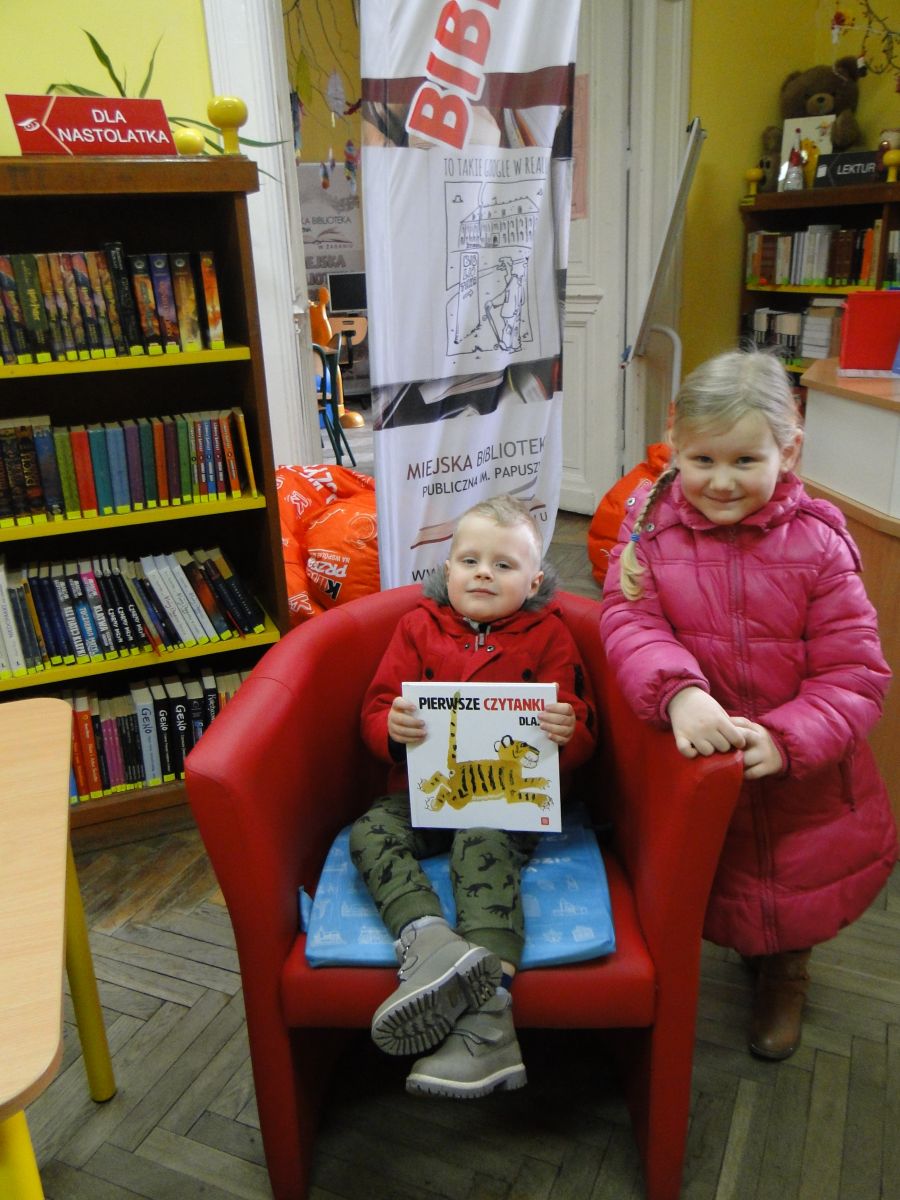 Na czerwonym fotelu siedzi trzyletni chłopczyk z otrzymaną książką, obok stoi dziewczynka. W tle baner Biblioteki i regały z książkami.