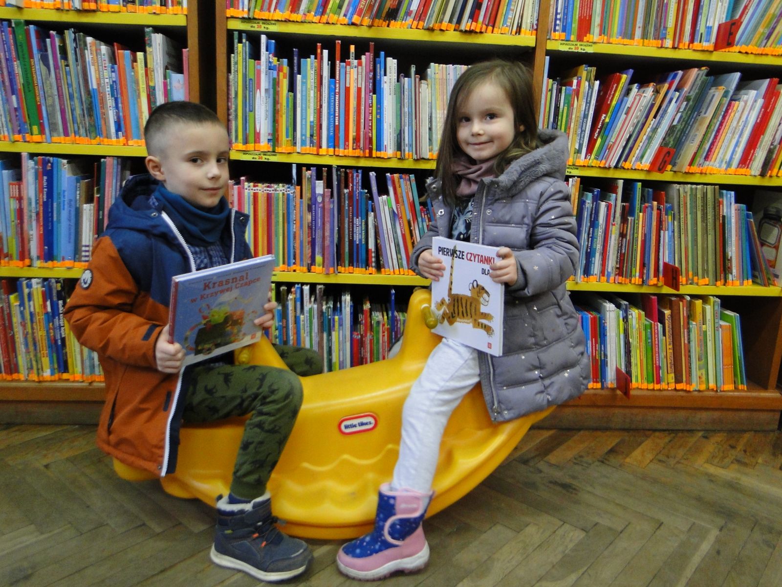 Na żółtej huśtawce siedzi dwoje dzieci, od lewej chłopiec, z prawej dziewczynka. Dzieci trzymają w rękach książki. W tle regały z książkami.