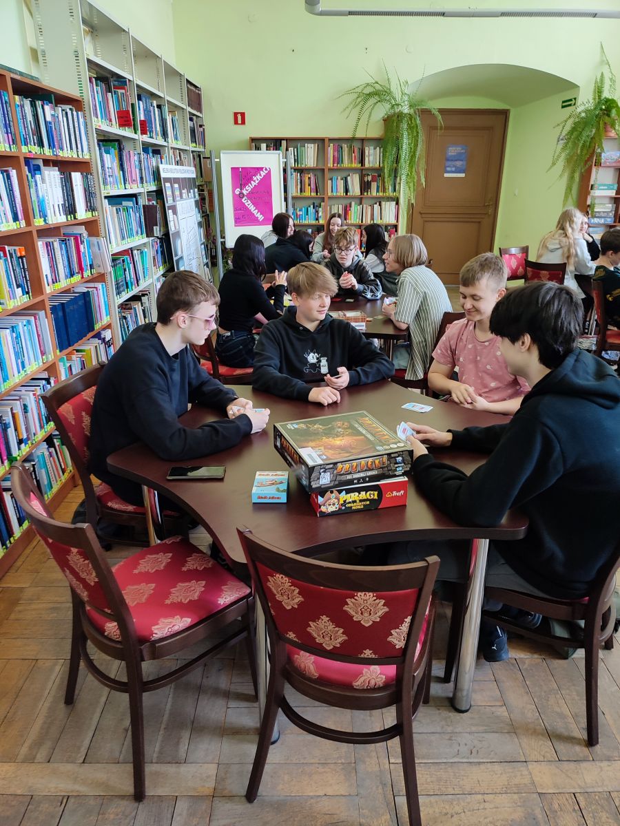 W Czytelni Ogólnej wokół stołów siedzą uczniowie, na stołach już rozłożone gry planszowe, uczniowie grają lub analizują instrukcję. W tle regały z książkami.