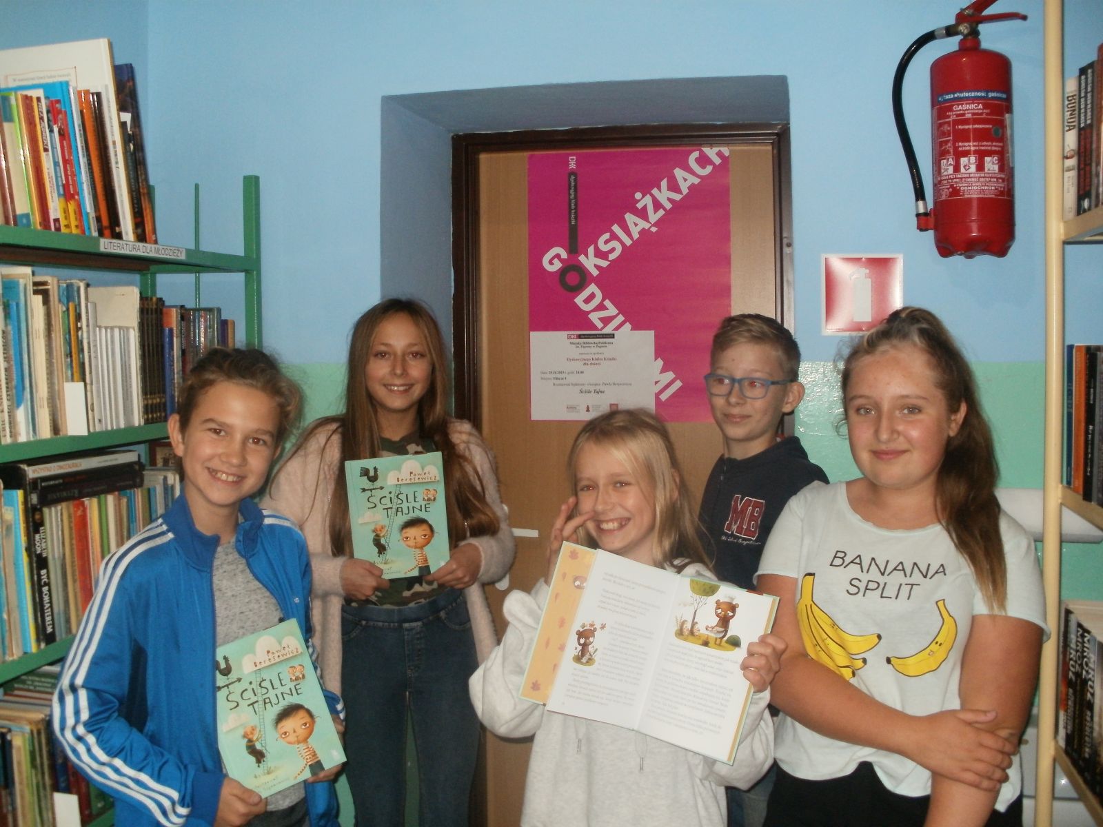 Zdjęcie. Pięcioro wesoło uśmiechniętych dzieci pozuje z przeczytaną książką na tle plakatu DKK Godzinami o książkach. W tle regały biblioteczne z książkami.