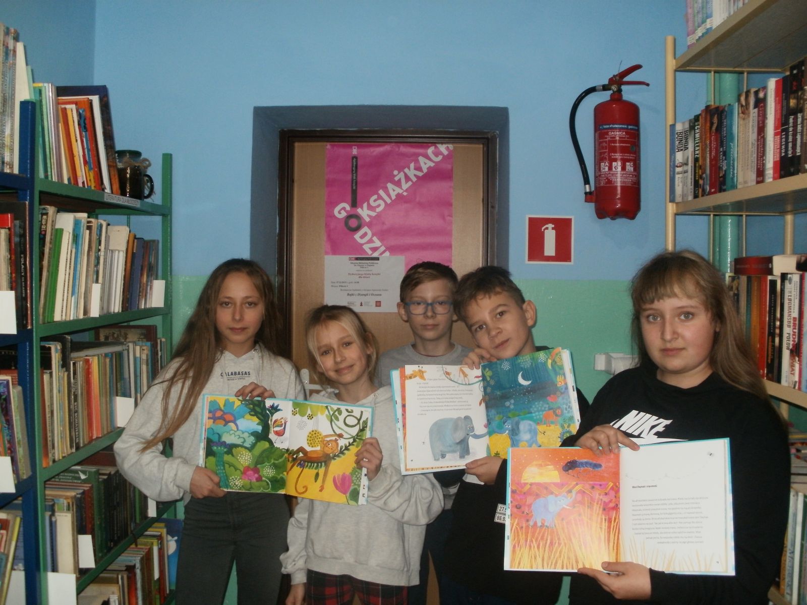 Zdjęcie. Pięcioro uśmiechniętych dzieci pozuje na tle plakatu Godzinami o Książkach. W tle regały biblioteczne z książkami.