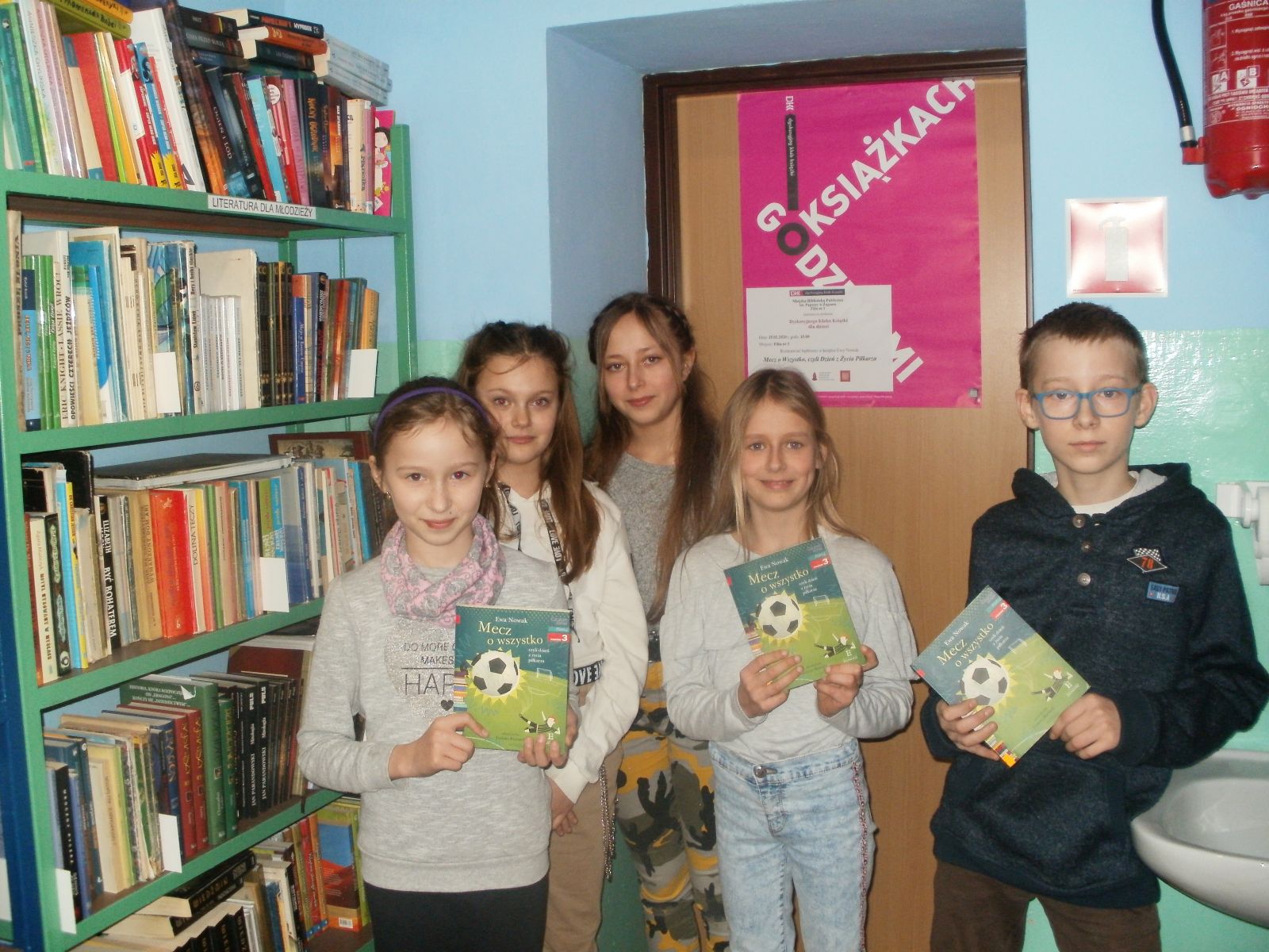 Zdjęcie. Na tle zielonego regału z książkami i plakatu Godzinami o książkach pozuje pięcioro uśmiechniętych dzieci trzymających przeczytaną książkę.